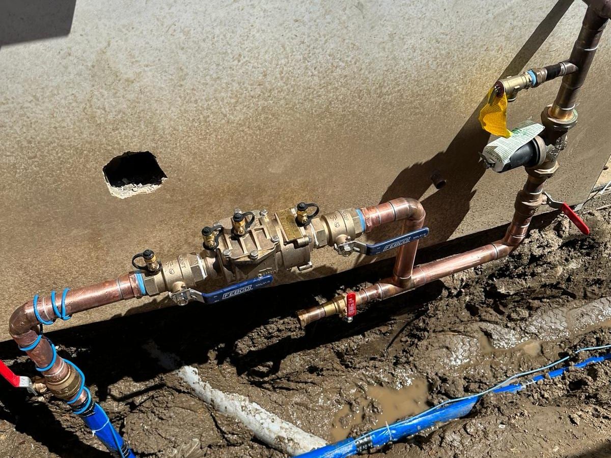 A brass backflow preventer with test cocks and blue-handled ball valves, piped in copper at the base of a wall