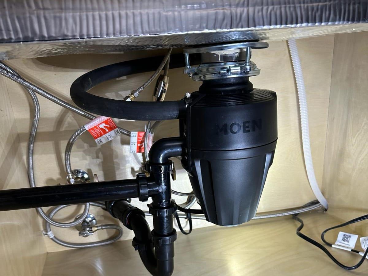 Under a kitchen sink, a black garbage disposal with its drain trap, the dishwasher hose and braided supply lines