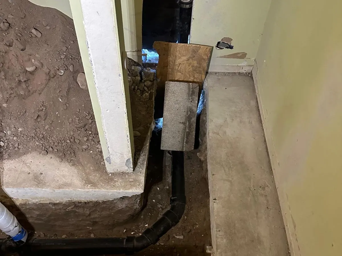 A trench cut through a concrete floor along a wall, with new black drain pipe laid in the soil