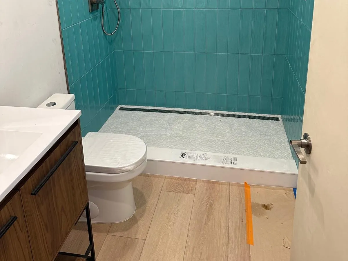 A new toilet beside a walnut vanity, facing an aqua tiled walk-in shower with a linear drain
