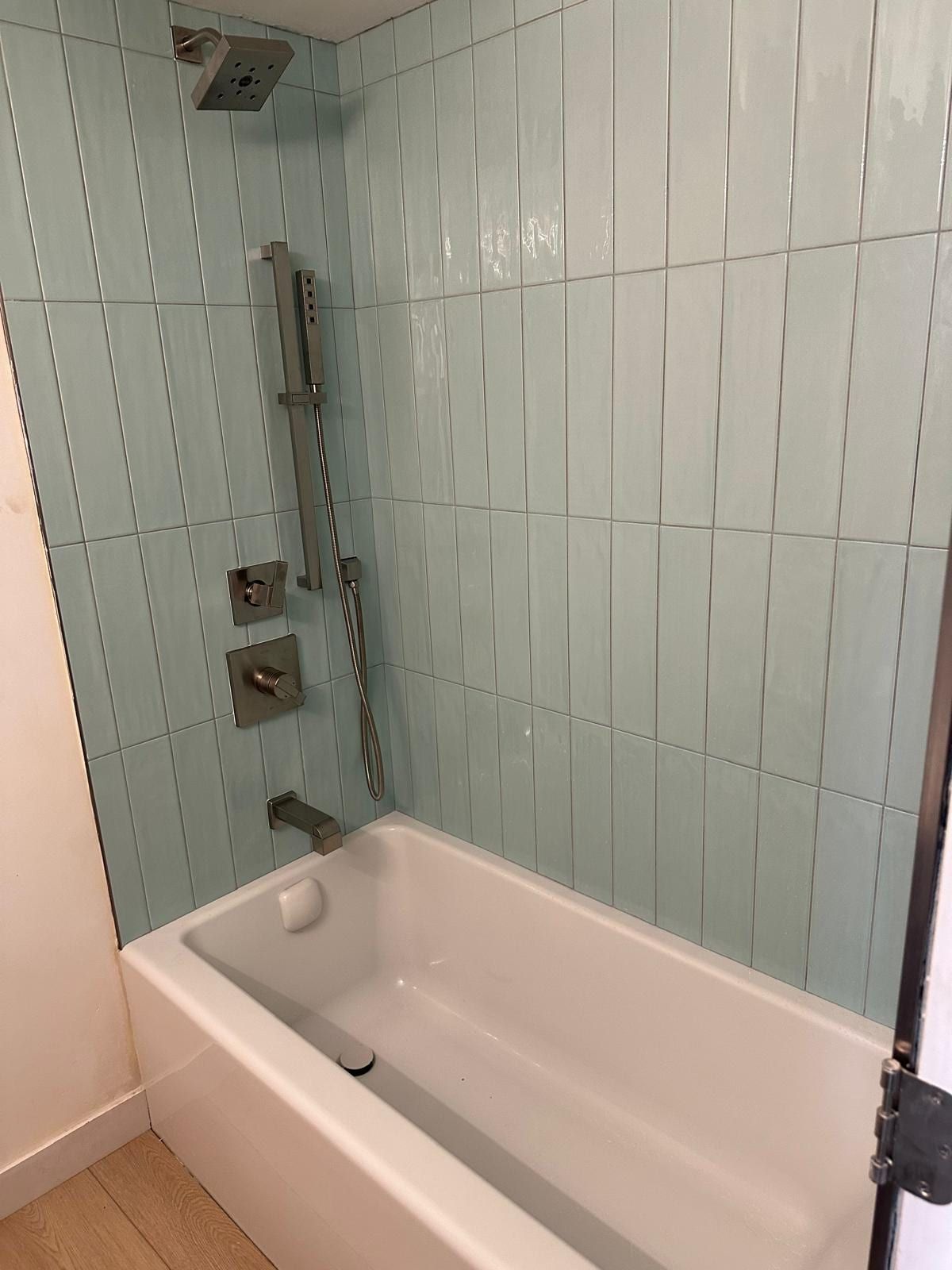 A tub and shower in pale aqua vertical tile with a square rain head, a slide bar with a hand shower and brushed nickel valve trim