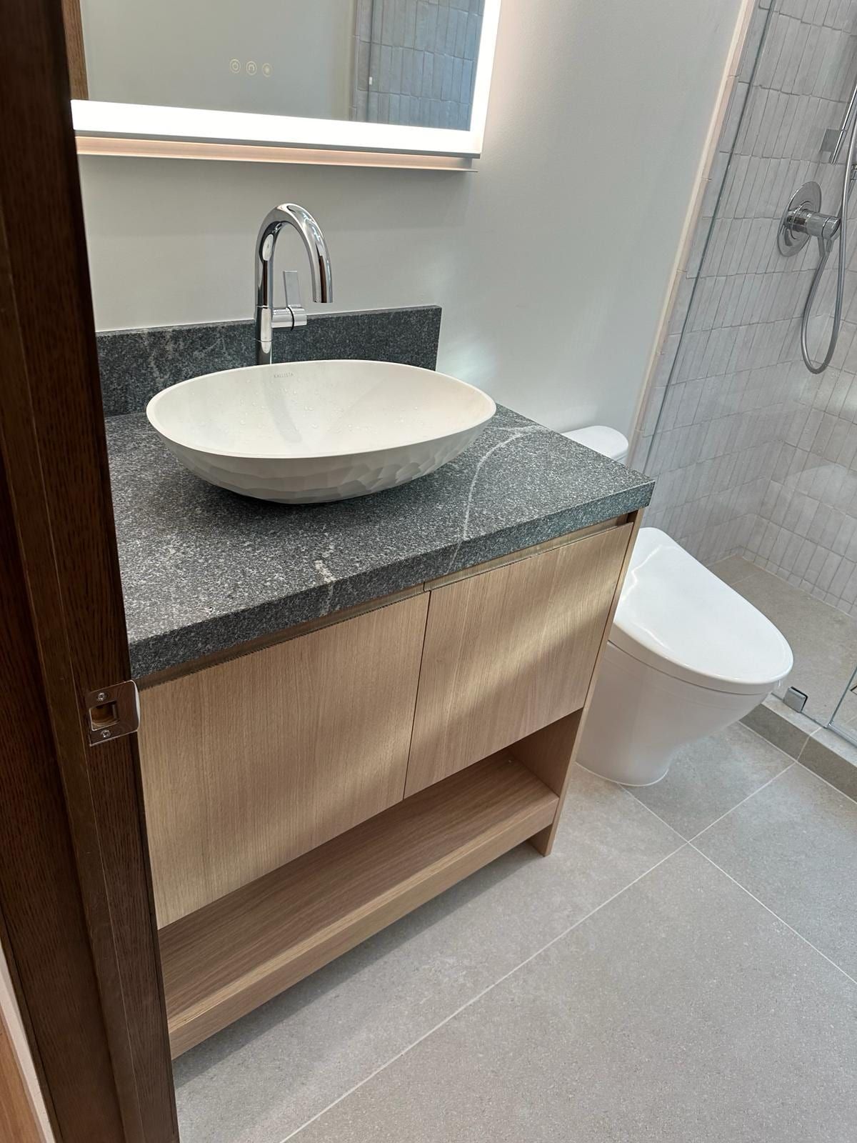 A light oak vanity with a dark stone top and a white vessel sink under a lighted mirror, with a toilet and a glass shower beyond